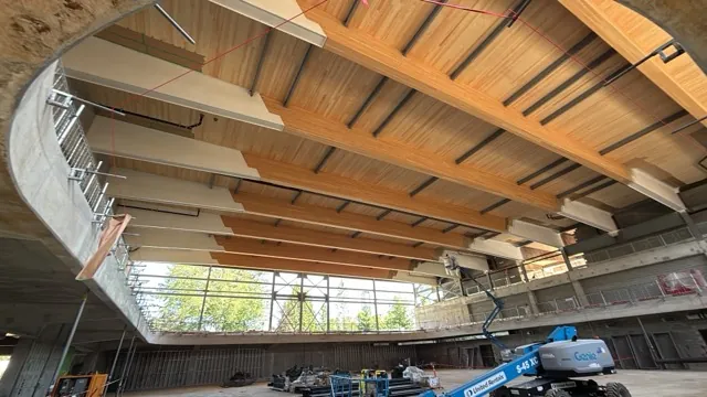 Mass timber roof of a large commercial hall — exposed glulam beams and cross-laminated timber panels finished with clear protective coating, with a boom lift on the floor below and clerestory glazing beyond