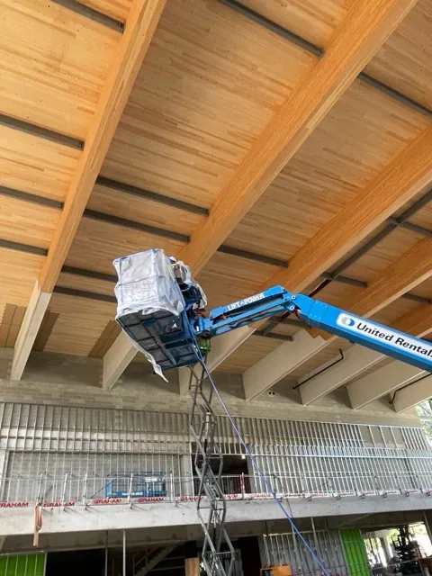 Boom lift raised to the underside of a mass timber roof while crew apply clear protective coating to exposed glulam beams and CLT ceiling panels